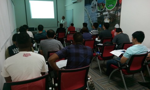 Classroom setting with a presenter and attendees facing a projector screen.
