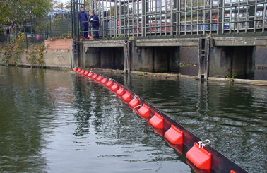 Floating barrier on water near a metal fence with two people observing.