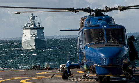 Blue helicopter on deck with a naval ship approaching in the background.
