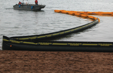 Boat deploying DESMI RO-CLEAN oil containment boom in water near shore.